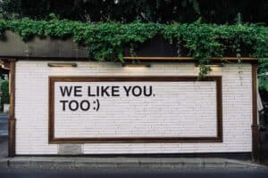 On top of a white brick wall it says, "We like you,too" Above, are green leaves and tree branches