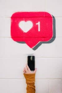 A hand holds up a smart phone against a white wall. Above, is a pink speech bubble with a white love heart and the number 1 next to each other, representing a reaction to a social media post.