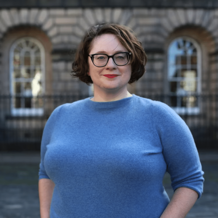 alt="A professional headshot of a woman standing in front of a historic Edinburgh building with arched windows. She is wearing a blue sweater, glasses, and has short brown hair styled in a bob cut. She is smiling warmly at the camera and wearing red lipstick. The architectural backdrop features classic stonework and arched windows typical of Edinburgh's historic buildings."