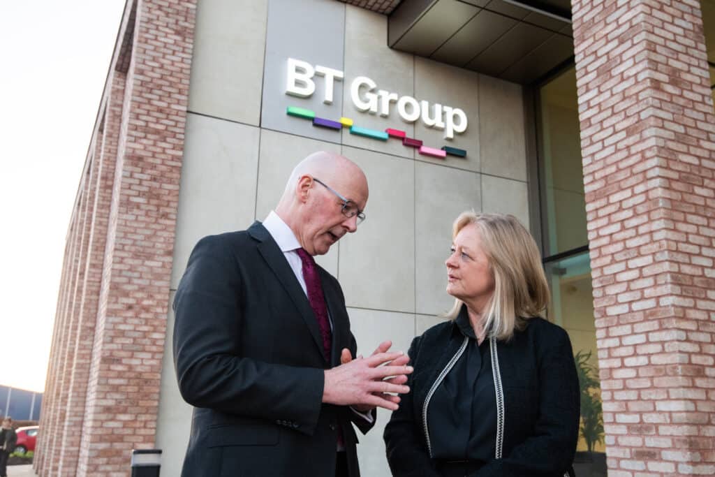 alt="First Minister John Swinney and BT Group CEO Allison Kirkby in conversation outside new BT Group Dundee office, standing beneath BT Group logo with its distinctive colorful bars. Both executives are engaged in professional discussion, with Swinney gesturing while speaking and Kirkby listening attentively. The building features modern architecture with exposed brick walls."