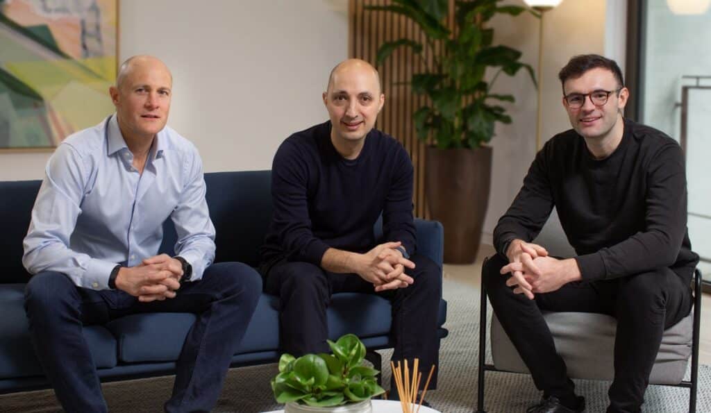 "Three men seated in a modern office setting. The man on the left wears a light blue dress shirt, the man in the middle wears a navy sweater, and the man on the right wears a black sweater and glasses. They are seated on navy blue furniture with indoor plants and artwork visible in the background. All three men are sitting in similar relaxed poses with their hands clasped."